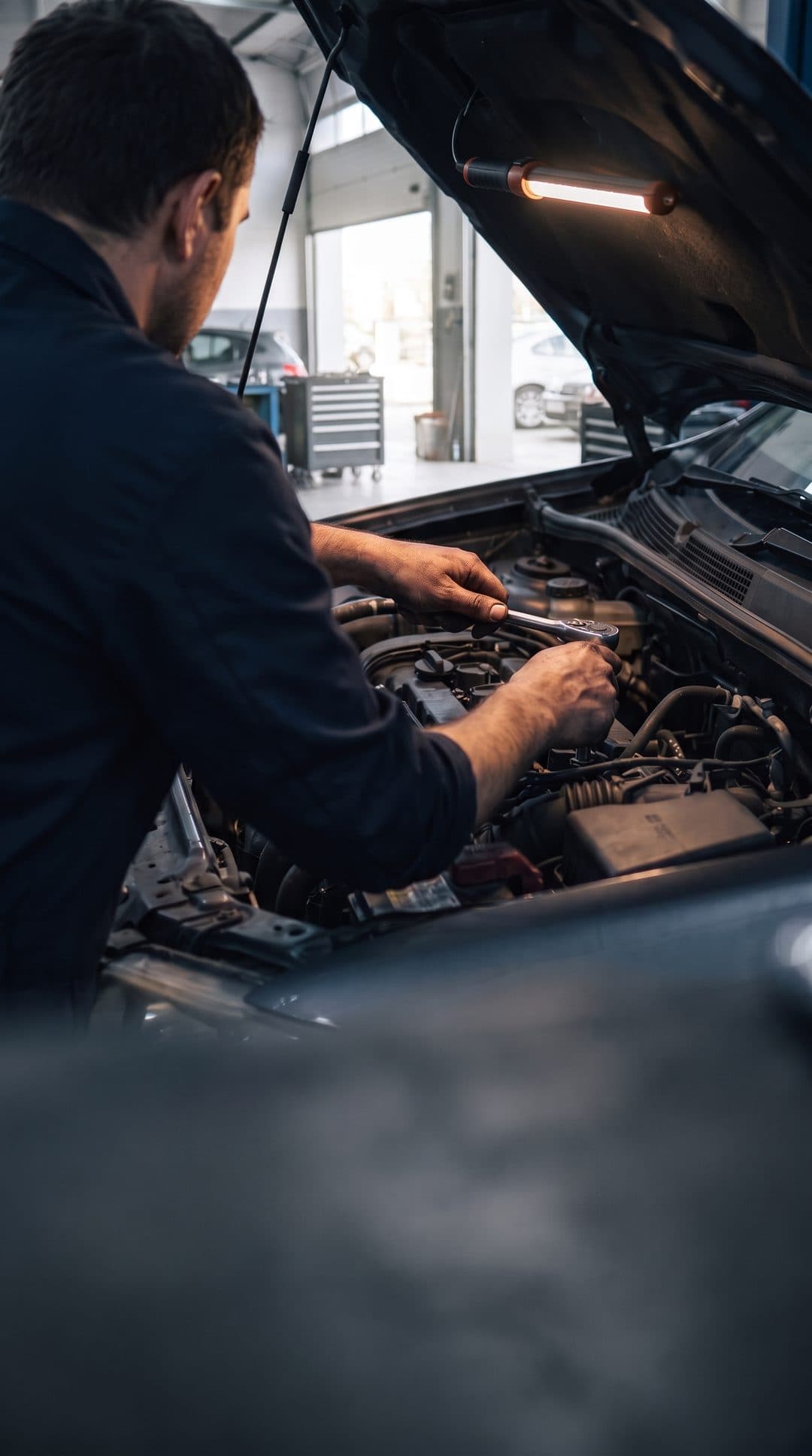 Mécanicien vu de dos, les mains dans le moteur d’une voiture capot ouvert, dans un atelier